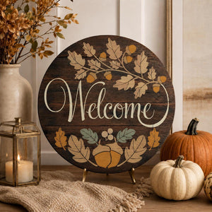 Round wooden 'Welcome' sign with autumn leaf and acorn illustrations, displayed on a tabletop with pumpkins, a lantern, and dried flowers in a home decor setting.