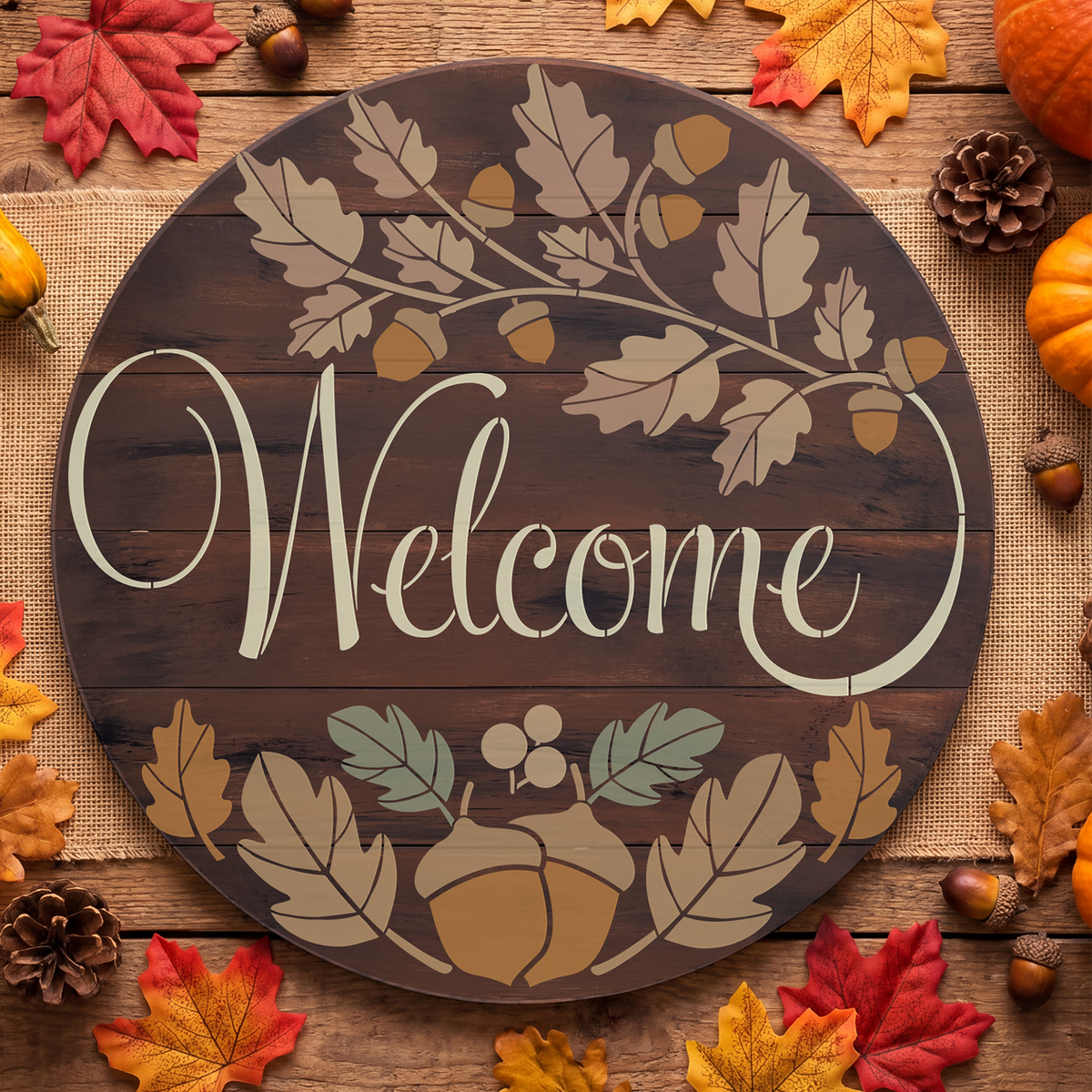 Round wooden 'Welcome' sign with autumn leaf and acorn motifs, styled on burlap with fall leaves, acorns, pinecones, and pumpkins around it.