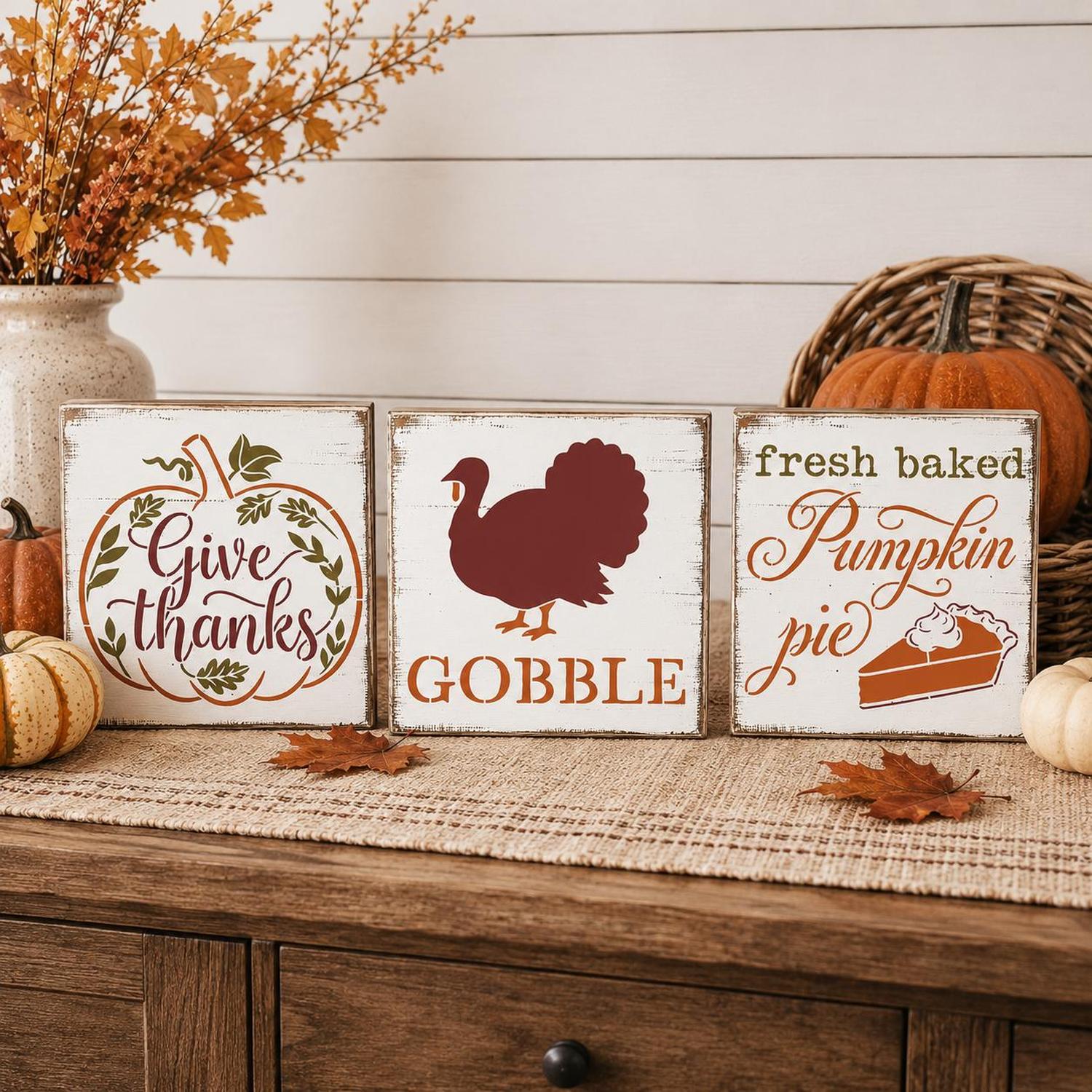 Three rustic white wooden sign plaques with autumn Thanksgiving-themed designs displayed on a burlap runner over a wooden cabinet. The signs read 'Give thanks' in a pumpkin border, 'GOBBLE' with a turkey silhouette, and 'fresh baked Pumpkin pie' with a pie slice graphic.