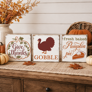 Three rustic white wooden sign plaques with autumn Thanksgiving-themed designs displayed on a burlap runner over a wooden cabinet. The signs read 'Give thanks' in a pumpkin border, 'GOBBLE' with a turkey silhouette, and 'fresh baked Pumpkin pie' with a pie slice graphic.