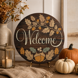Round wooden 'Welcome' sign with autumn leaf and acorn illustrations, displayed on a tabletop with pumpkins, a lantern, and dried flowers in a home decor setting.